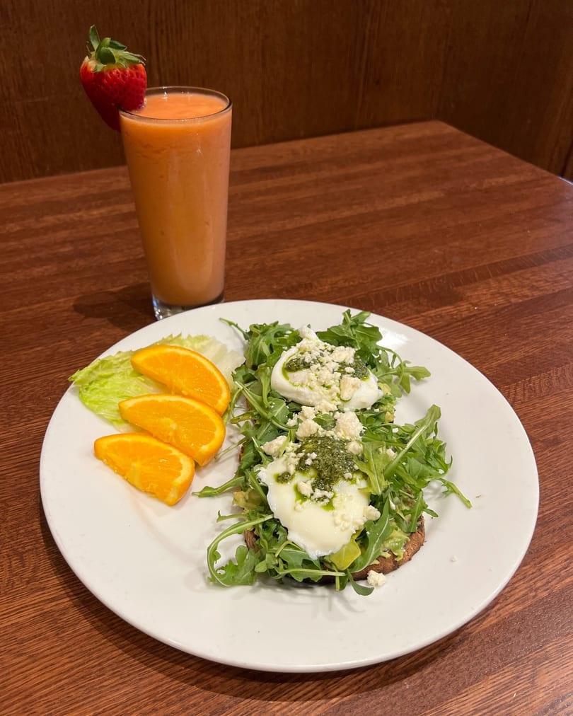 Avocado toast with poached eggs, pesto, feta, arugula, orange slices, and a strawberry smoothie.