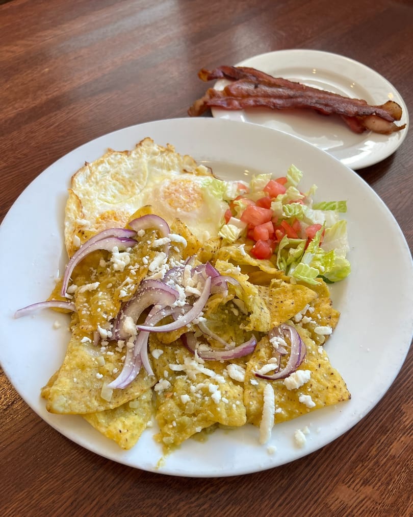 Chilaquiles with green sauce, fried eggs, red onions, cheese, lettuce, tomatoes, and bacon on the side.