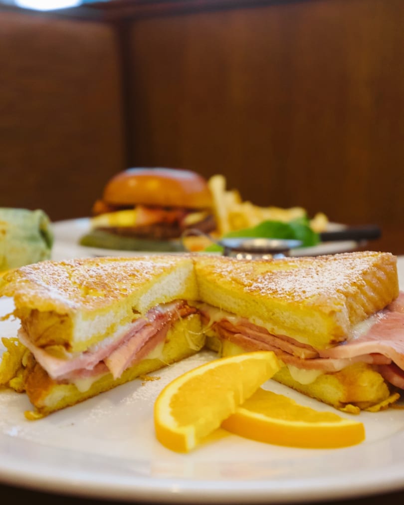 Close-up of a Monte Cristo sandwich with powdered sugar and orange slices, with other dishes blurred in the background.